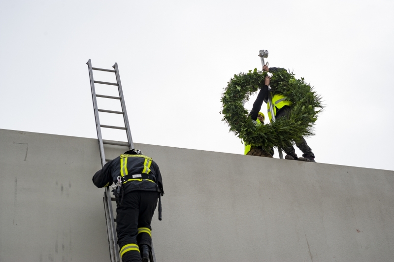 Ugunsdzēsējs kāp pa kāpnēm uz jumta, kur cilvēks tur lielu vainagu