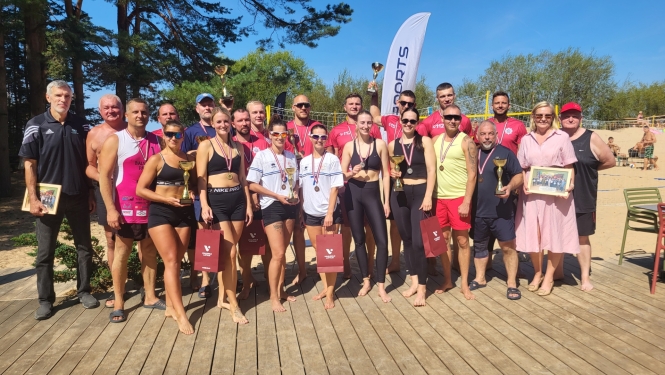 Plaša pludmales volejbola turnīra dalībnieku kopbilde saulainā dienā uz koka klāja. Daļa dalībnieku ir sporta tērpos ar medaļām kaklā un kausiem rokās. Fonā redzams smilšu volejbola laukums un mežs.
