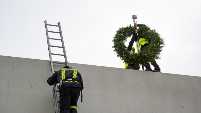 Ugunsdzēsējs kāp pa kāpnēm uz jumta, kur cilvēks tur lielu vainagu