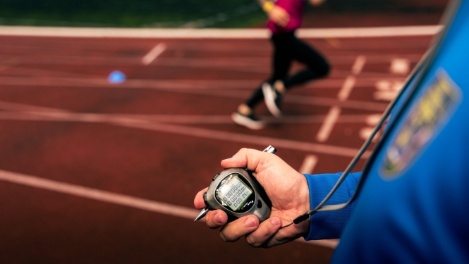Priekšplānā hronometrs cilvēka rokā, tālākā plānā - cilvēks skrien pa sarkanu gumijas celiņu