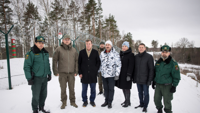 Priekšplānā cilvēki, aizmugurē žogs un apsnidzis mežs