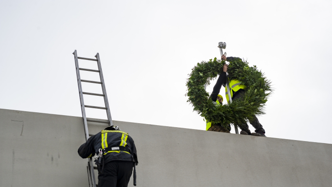 Ugunsdzēsējs kāp pa kāpnēm uz jumta, kur cilvēks tur lielu vainagu