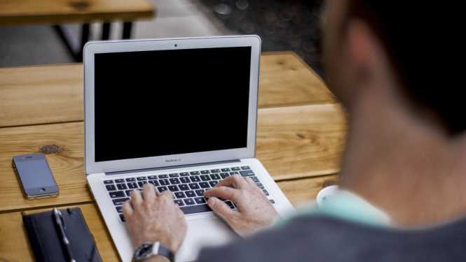 cilvēks galda, uz kura ir portatīvais dators, telefons un piezīmju klade ar pildspalvu