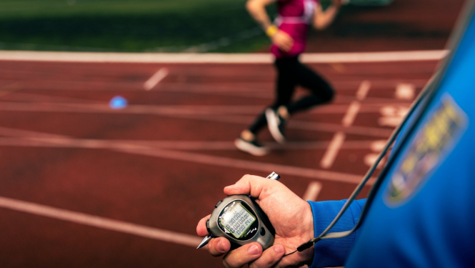 Priekšplānā hronometrs cilvēka rokā, tālākā plānā - cilvēks skrien pa sarkanu gumijas celiņu
