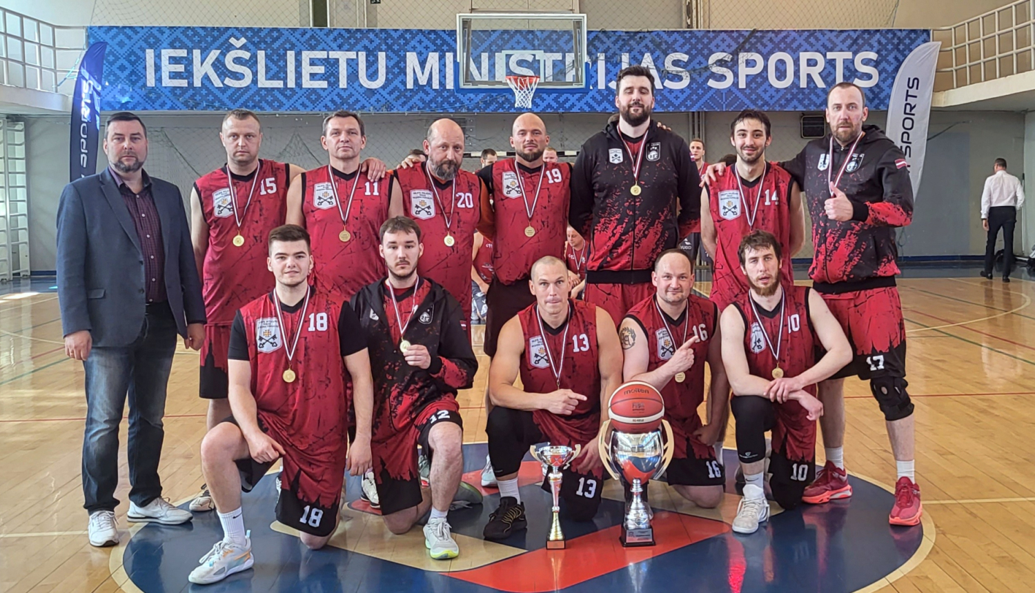 2026.gada Iekšlietu ministrijas čempionāta čempioni basketbolā – Valsts policijas Rīgas reģiona pārvaldes komanda sporta zālē, spēlētājiem kaklā zelta medaļas, priekšā ceļojošais čempionu kauss un pirmās vietas ieguvēju kauss.