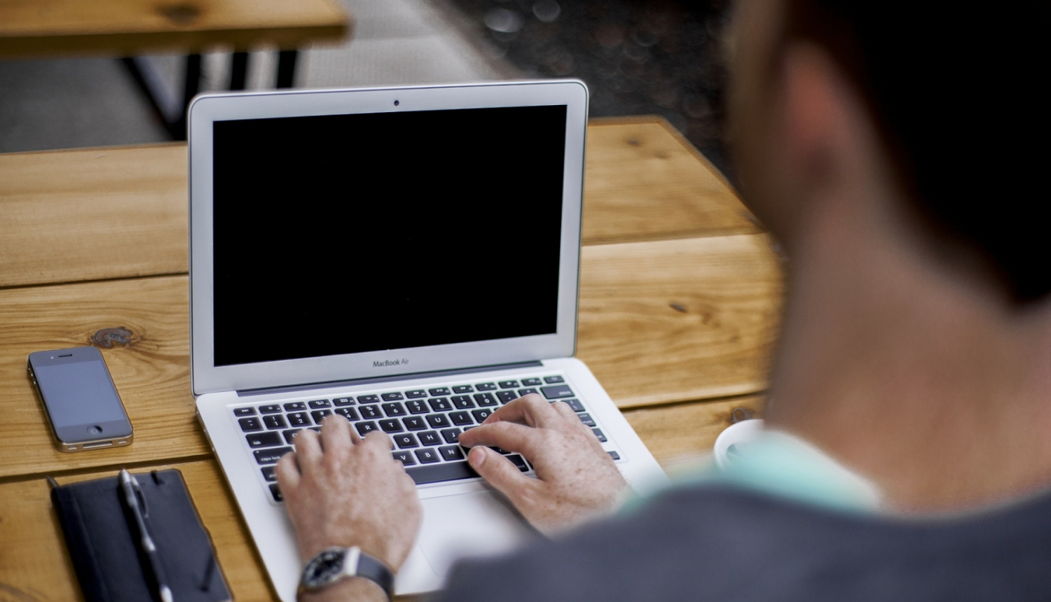 cilvēks galda, uz kura ir portatīvais dators, telefons un piezīmju klade ar pildspalvu