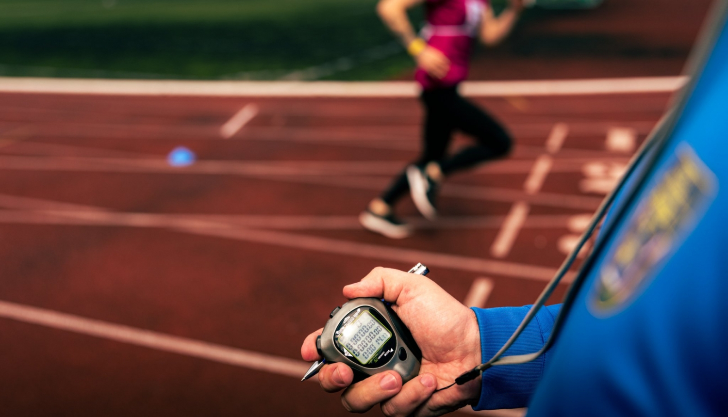 Priekšplānā hronometrs cilvēka rokā, tālākā plānā - cilvēks skrien pa sarkanu gumijas celiņu